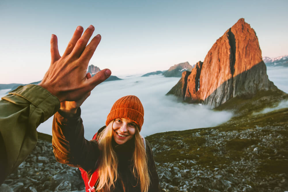A girl hiking. She's standing on top of a moutain with a stunning view, high-fiving a hand that just appears in the frame. In the background there's clouds and mountain peaks. 