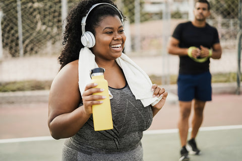A woman of colour with dark, curly hair in exercise clothes. She's wearing headphones and a towel around her neck, while holding a drink bottle. She's ready to go! 